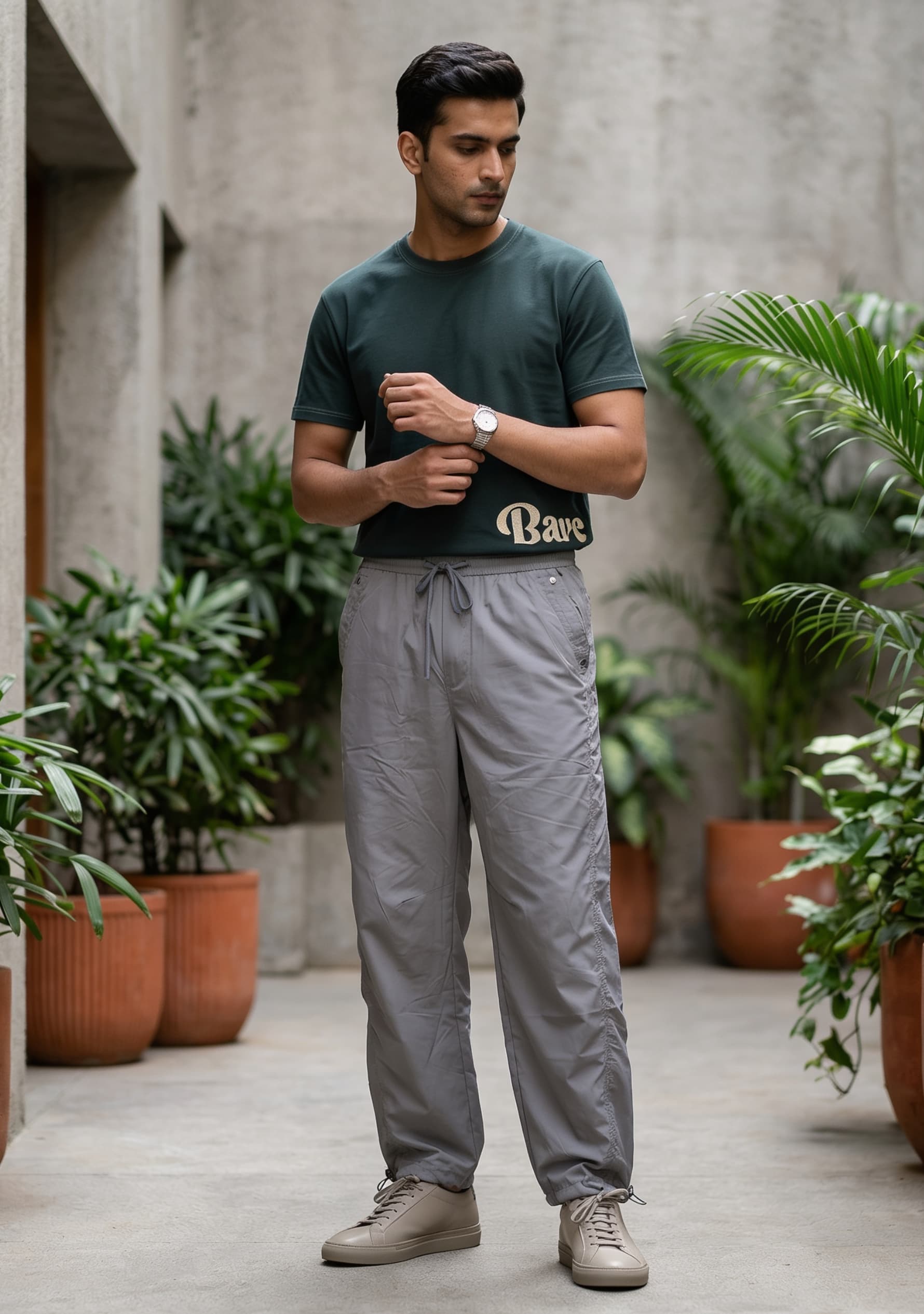 Man wearing a green t-shirt and gray pants standing in an outdoor setting with plants.